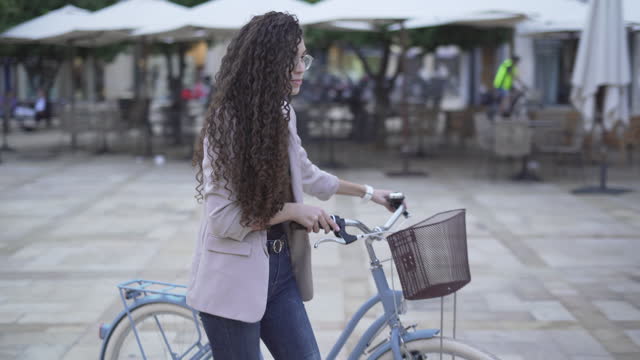 Charming Young Female Model In Glasses With Long Curly Hair Walking With A Bicycle Outdoor. medium shot