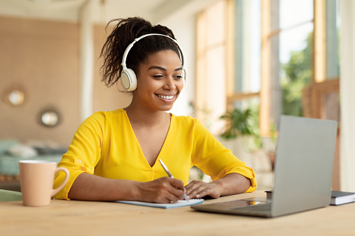 Smiling black lady in wireless headphones working on laptop and taking notes in paper notebook, watching webinar, sitting at desk at home, free copy space