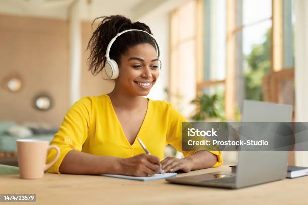 Smiling Black Lady In Wireless Headphones Working On Laptop And Taking Notes In Paper Notebook Watching Webinar Stock Photo - Download Image Now