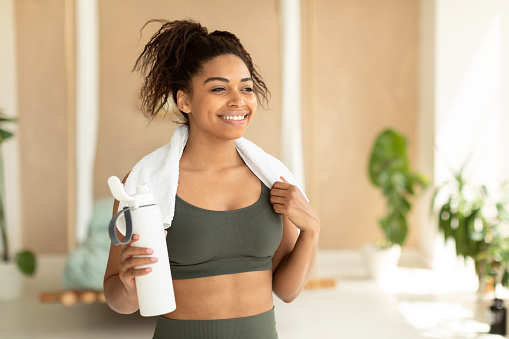 Portrait of happy black woman in sportswear and white towel on neck holding bottle with fresh water, having rest after domestic workout, free copy space