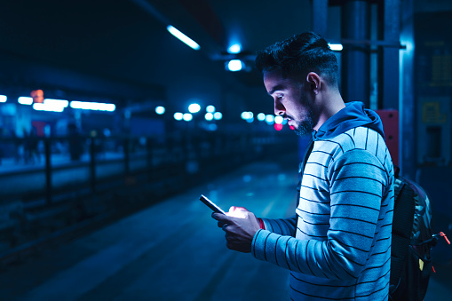 Indian young man with backpack standing on railway station platform and using a smartphone while waiting for the train at night.