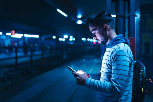 Young man using a smartphone while waiting for the train. Indian young man with backpack standing on railway station platform and using a smartphone while waiting for the train at night. subway platform stock pictures, royalty-free photos & images