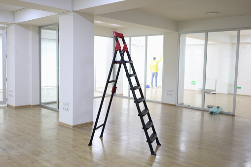 Metal ladder standing in center of empty office, worker repairing wall in background. Office renovation and improvement concept.