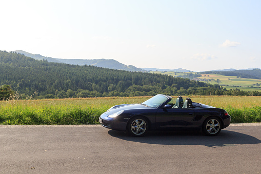 Gersfeld, Germany - July 23, 2021: Blue roadster Porsche Boxster 986 with Rhön Mountains panorama. The car is a mid-engine two-seater sports car manufactured by Porsche.
