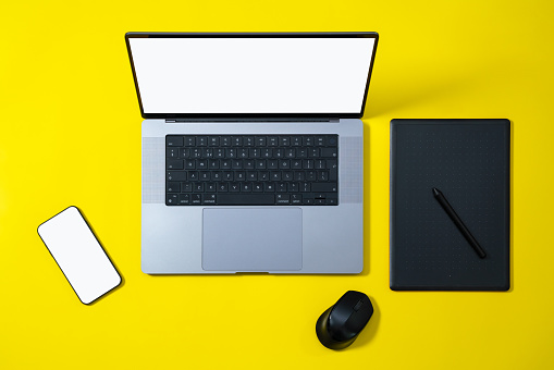 Business workspace flat lay. Modern laptop, phone with blank white screen and graphics tablet and computer mouse on yellow background. High angle view.