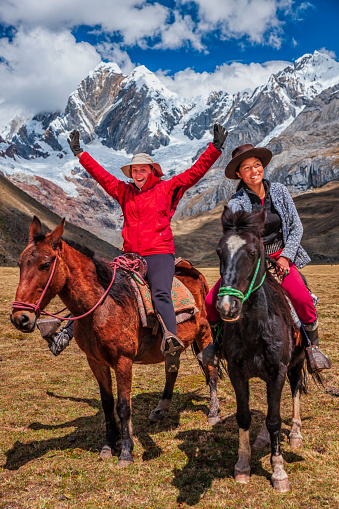 Young girl riding a horse with a young female tourist, Peruvian Andes, South America. Andes Mountain Range is located in South America, running north to south along the western coast of the continent.It is a continual range of highlands along the western coast of South America.The Andes extend from north to south through seven South American countries: Argentina, Bolivia, Chile, Colombia, Ecuador, Peru, and Venezuela, and is the longest continental mountain range in the world.