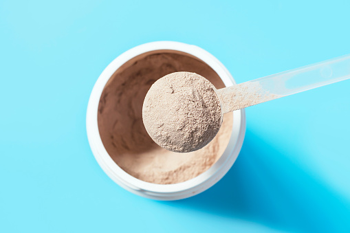 Chocolate protein powder in a scoop against a plastic jar on a blue background.
