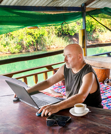 Working remotely on his computer at Si Phan Don,a man traveling the world,sits on a balcony at a rural guesthouse,overlooking the Mekong river,with a cup of coffee,in a beautiful,relaxed setting.