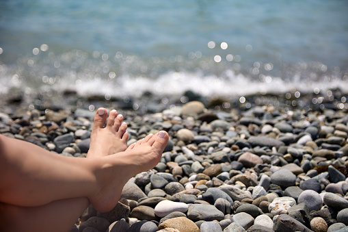 Close-up of a woman's feet resting on a rocky shore against the backdrop of splashing waves. Girl relaxes, relaxes on the beach on a vacation by the sea