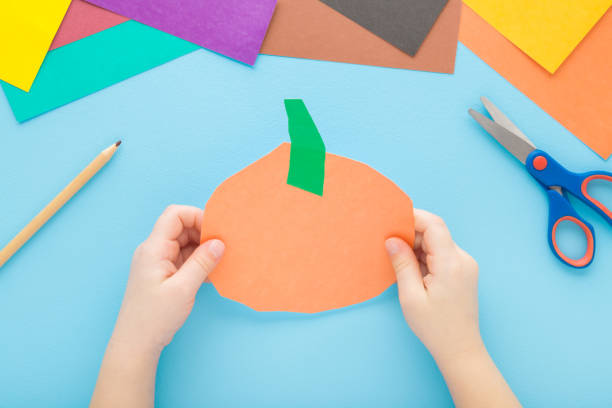 istock Little child hands holding orange pumpkin shape from paper on light blue table background. Pastel color. Closeup. Point of view shot. Toddler development. Top down view. 1474783702
