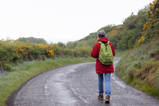 Rear view shot of a senior woman on a hiking staycation in Dumfries and Galloway, Scotland. She is walking along a road with a backpack on.