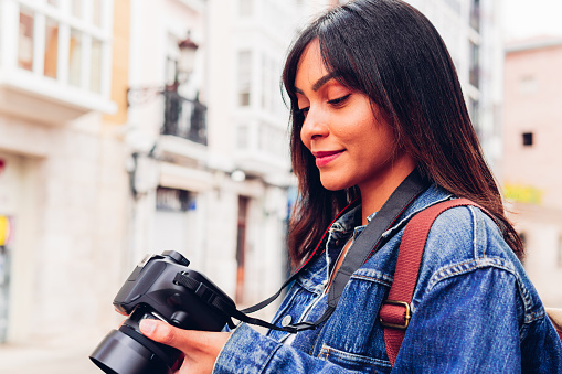 Smiling young Hispanic lady in denim jacket checking photos on camera while standing in street of old town
