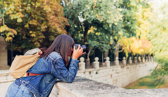 Side view of woman in denim outfit with backpack leaning on stone border of bridge in sunny park and taking picture of nature