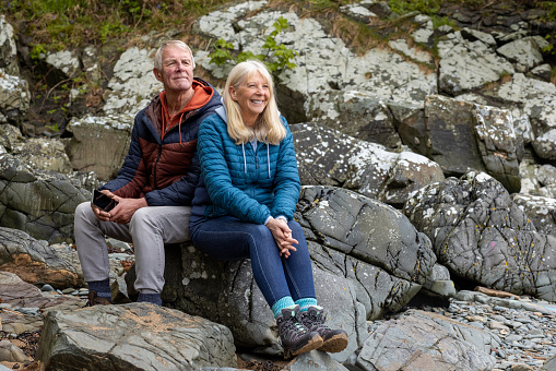 Senior couple on a hiking staycation in Dumfries and Galloway, Scotland. They are at the coast on a beach, taking a break while sitting on rocks.