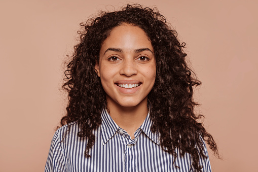 Close up portrait of young happy smiling multiracial woman face, with curly hair, wears striped shirt, posing at studio isolated over beige wall background.