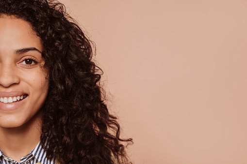 Half face portrait of a young multiracial beautiful smiling woman with curly hair, isolated over beige background. Copy space place for your advertising content.