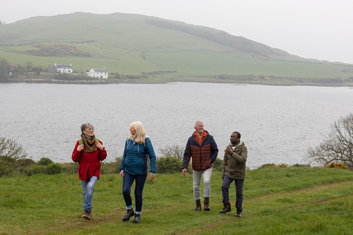 Senior friends on a hiking staycation in Dumfries and Galloway, Scotland. They are walking through a grassy rural area with a lake visible in the distance behind them.