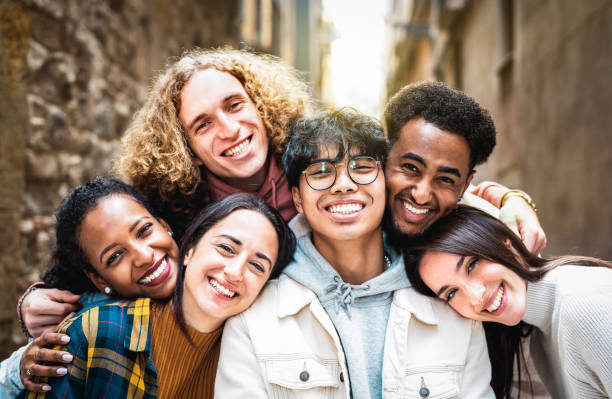 Multi racial guys and girls taking selfie outdoors with backlight - Happy life style friendship concept on young multiracial best friends having fun day together in Barcelona city - Warm vivid filter stock photo
