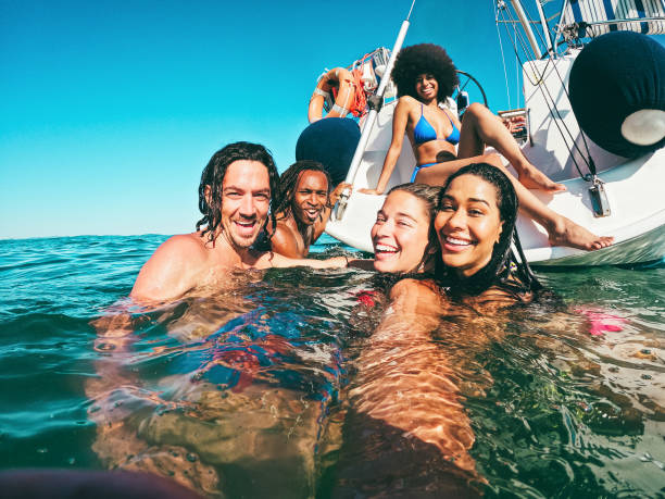 Happy multiracial friends doing selfie swimming in the sea with sail boat in background - Soft focus on center girl face Happy multiracial friends doing selfie swimming in the sea with sail boat in background - Soft focus on center girl face yacht selfie stock pictures, royalty-free photos & images