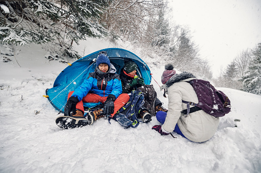 Three teenage kids are hiking in European Alps on a winter day. They are sitting by a tent in the forest.\nBeautiful snowy winter day. \nShot with Canon R5