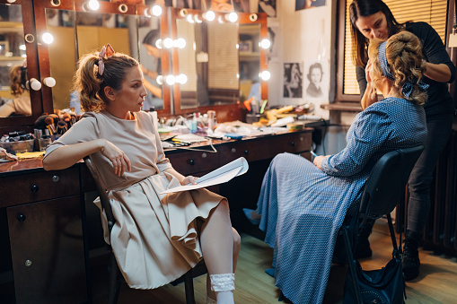 An actress who has already done her make-up and hair for the play is sitting and reading the text, while make-up artist is doing her senior co-star's makeup