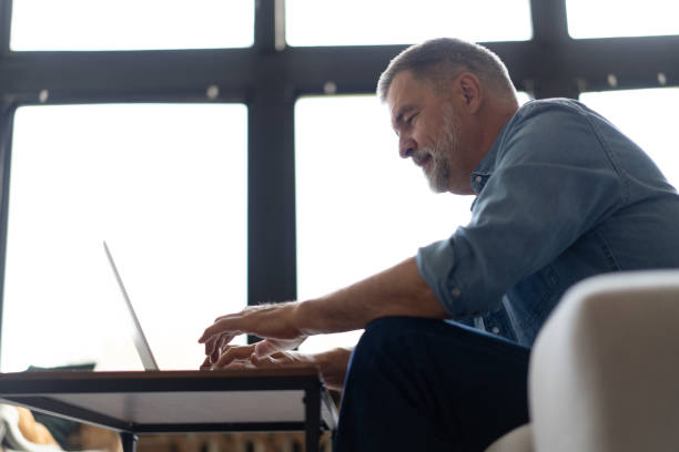 istock Senior man in casual clothing using laptop and smiling while sitting on the sofa, working from home. 1474802513