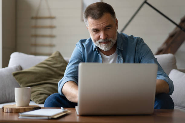 istock Senior man in casual clothing using laptop and smiling while sitting on the sofa, working from home. 1474802514