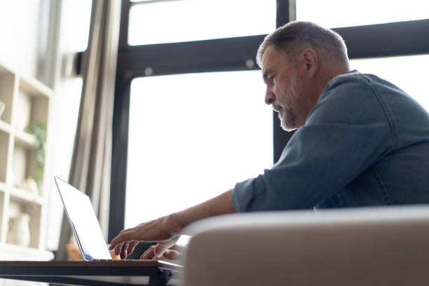 istock Senior man in casual clothing using laptop and smiling while sitting on the sofa, working from home. 1474802516