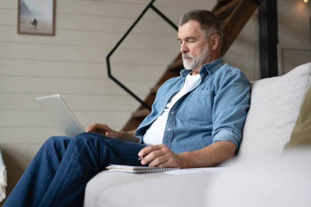 istock Senior man in casual clothing using laptop and smiling while sitting on the sofa, working from home. 1474802531