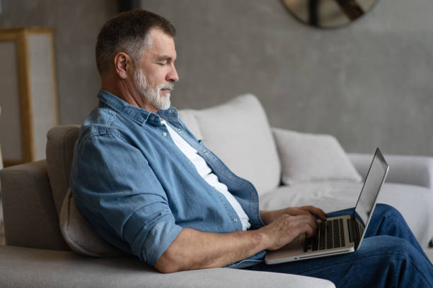 istock Senior man in casual clothing using laptop and smiling while sitting on the sofa, working from home. 1474802537
