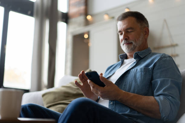 istock Happy smiling senior man using smartphone device while sitting on sofa at home 1474802612