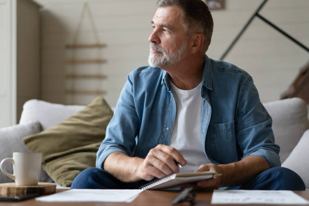 istock Senior man taking notes in notebook while sitting on the couch, gray-haired elderly man writing thoughts in notebook 1474802670