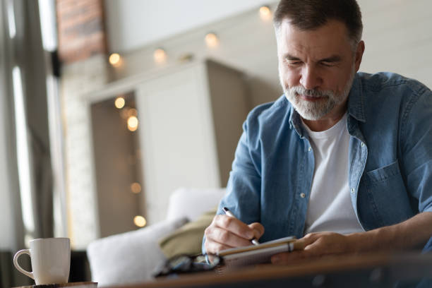 istock Senior man taking notes in notebook while sitting on the couch, gray-haired elderly man writing thoughts in notebook 1474802671