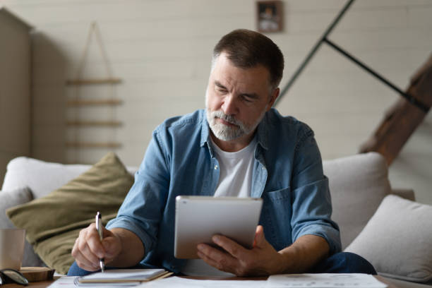 istock Cheerful senior man in casual clothing using digital tablet while sitting on the sofa at home. 1474802742