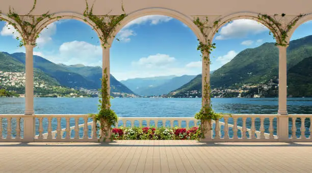 Beautiful view from the terrace over Lake Como and the mountains. Terrace with columns and balustrade of a Mediterranean palazzo on a sunny day. Flowers and ivy in the foreground.