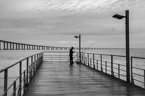 Old man fishing on Pier on the banks of the Tagus River next to the Vasco da Gama bridge in Lisbon