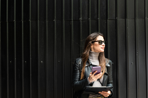 Cool Rock Businesswoman dressed in leather jacket small internet company owner standing outdoor corporate building. Female business person with sunglasses on city street near office building