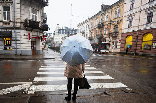 Ivano-Frankivsk, Ukraine - March, 2023: Back of woman with umbrella stand near pedestrian croos in rain street.