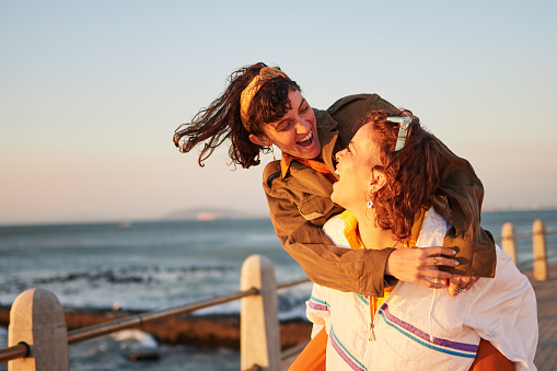 Women, gay couple and piggyback by the ocean coast with smile for happy relationship and fun pride in the outdoors. Lesbian woman on back ride with partner laughing in joy and happiness by the beach