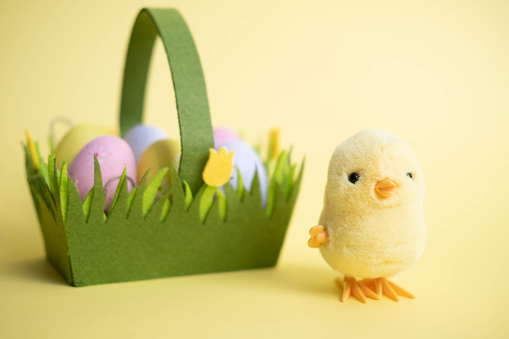 Easter card. a small yellow toy chicken, next to a green basket with colorful eggs, on a yellow background. We are celebrating Easter