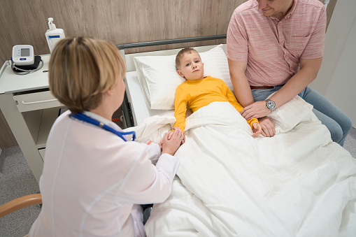 Waist up portrait of attractive little boy is lying on the medical bed while doctor and his father is sitting hear in the ward