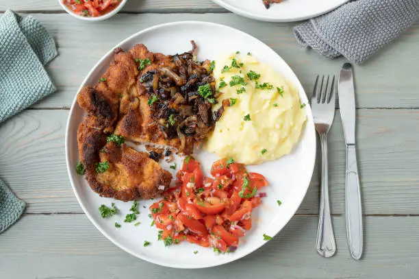 Delicious homemade german dinner or lunch with pan fried breaded pork schnitzel. Served with fried onions, mashed potatoes and tomato salad on a plate with cutlery and napkins. Served on light wooden table from above