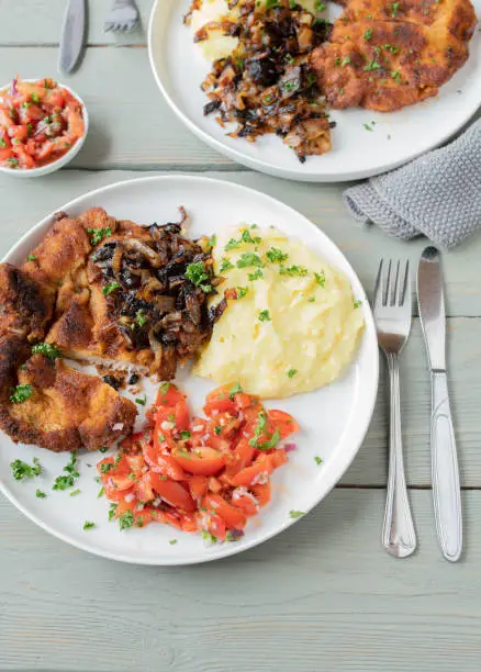 Homemade dinner or lunch, traditional cuisine with a fresh breaded pork schnitzel. Served with roasted onions, mashed potatoes and tomato salad on a plate with cutlery on light wooden table background from above.