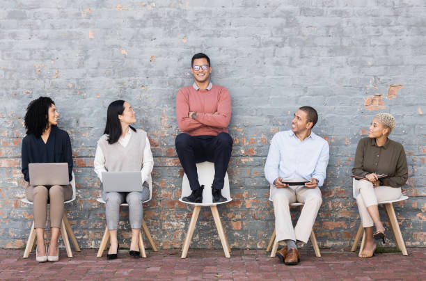 istock Creative business people, waiting room and man standing out against a brick wall for interview, meeting or opportunity. Group of employee interns with technology looking at candidate for startup 1474830825