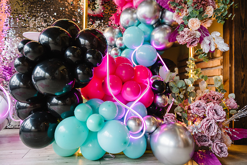 Luxury arch is decorated with flowers. Silver wall for party time. Light blurred background for birthday. Photo zone with black lights, silver, pink, blue balloons, disco style, led strip. Details.