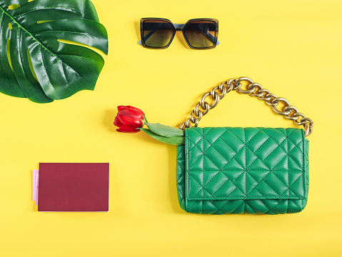 Stylish women's trendy green quilted bag with a large gold chain, red tulip, passport and manster leaf on a yellow background, flat lay closeup. The concept of fashionable accessories, travel, banner.