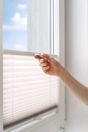 cropped shot of female hand pull and close pleated jalousie on window in living room, controlling lighting range, protection of sunlight, closeup