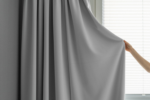 cropped shot of female hold grey fabric curtain and close window for protection from sunlight in living room with modern style, privacy and comfort concept