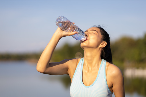 Thirsty Asian female athlete drinking water after exercising outdoors in park.