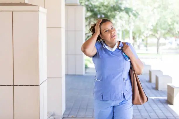 An emotionally and physically exhausted mid adult female nurse rubs her head as she leaves the hospital.